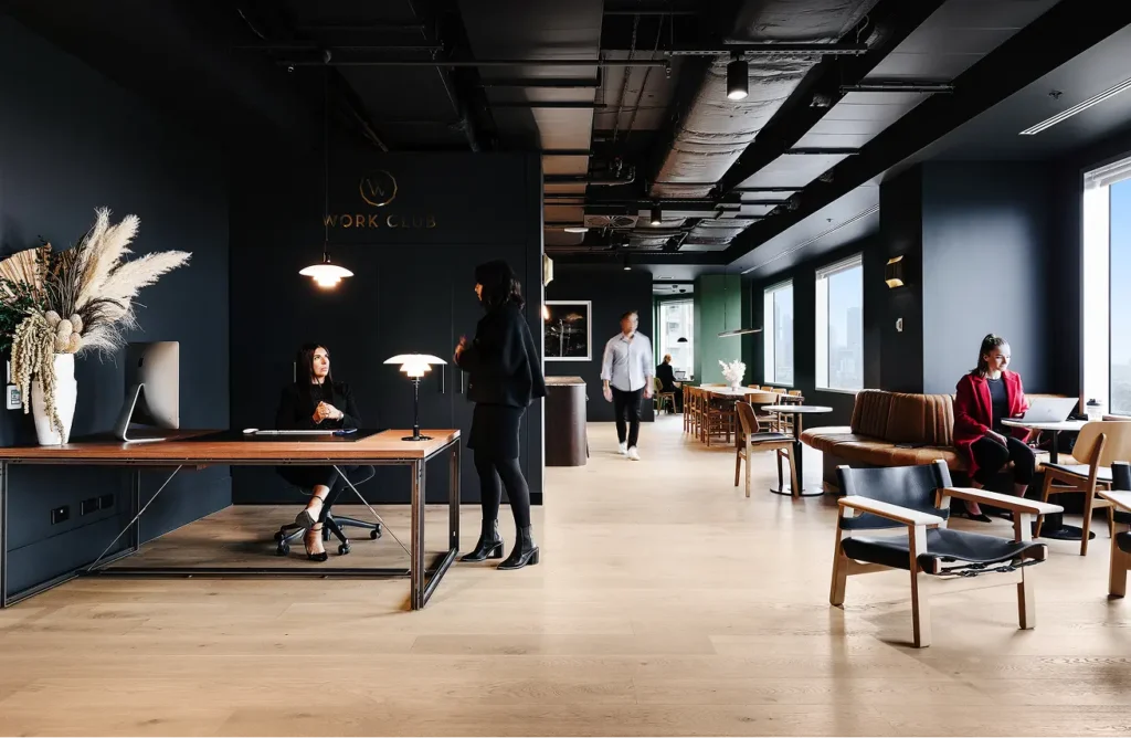 Professionals work and interact inside a modern, dark-walled co-working lounge featuring wooden floors, large windows, sleek seating, and a reception area with a wall reading "WORK CLUB".