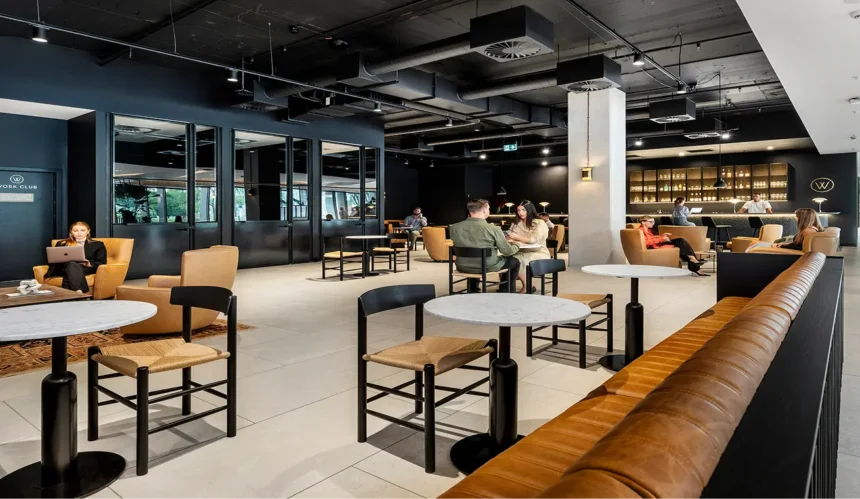 People converse and work at tables and leather armchairs inside a modern co-working lounge with dark walls and exposed ceiling ductwork. Visible text includes: (W) WORK CLUB FIRE SAFETY DOOR DO NOT OBSTRUCT DO NOT KEEP OPEN (W)