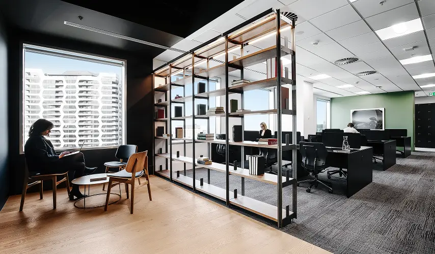 A tall, open-frame metal bookshelf divides a modern office interior, separating a dark-walled seating area with a city-view window from a bright, carpeted workspace filled with desks.