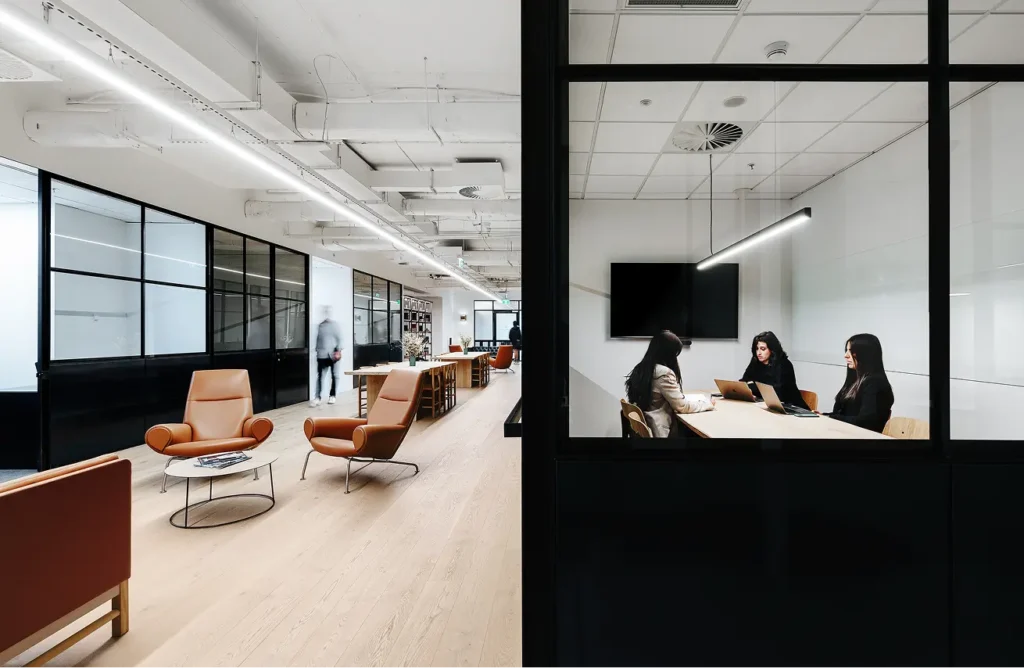 Three office workers sit around a conference table inside a glass-walled meeting room, adjacent to a modern open office hallway featuring leather armchairs and light wood flooring.