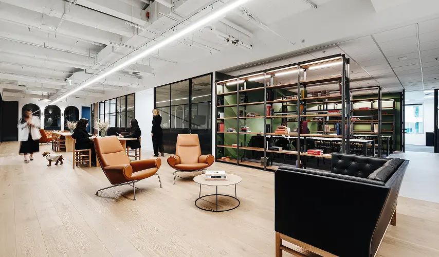 People sit, walk, and work throughout a spacious, modern office lounge furnished with tan leather armchairs, a black sofa, tall metal bookshelf partitions, and glass-partitioned meeting rooms.