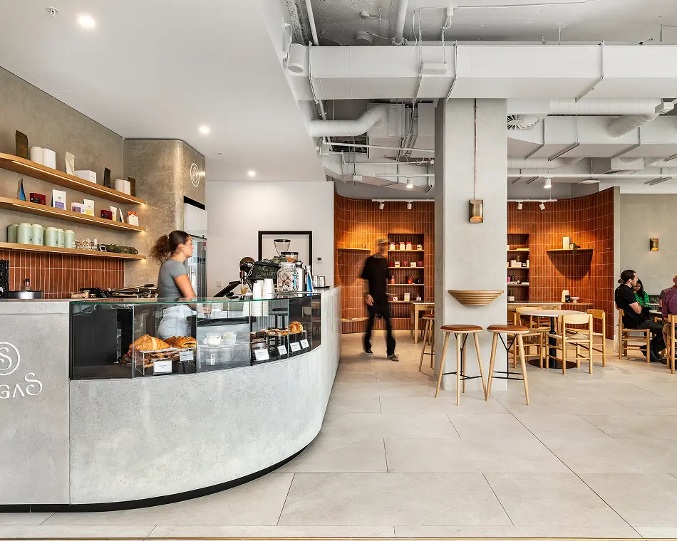 A barista stands behind a curved concrete service counter with a glass pastry case inside a modern cafe featuring terracotta tiled walls, exposed ceiling piping, and minimalist wooden seating. Text on the counter reads: "SGAS"