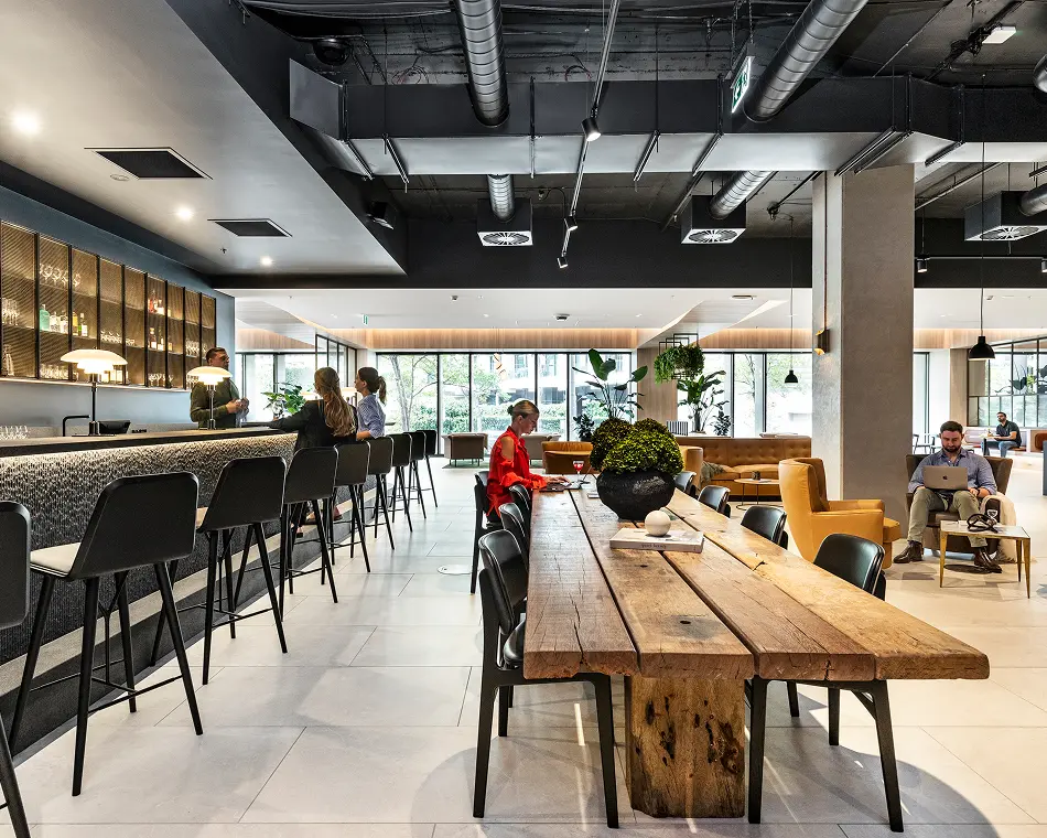 People sit, work on laptops, and socialize around a long wooden communal table and a sleek bar inside a modern lounge with industrial black ceiling pipes and large windows.