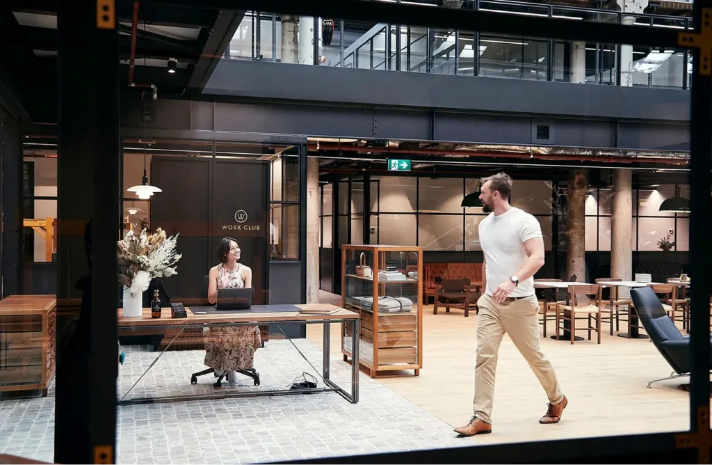 A man in a white t-shirt walks through a spacious modern co-working office while a woman waves from a reception desk. Text on a glass wall reads "W WORK CLUB".