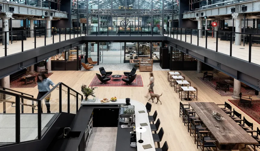 A woman descends stairs while another walks a dog across a spacious, two-story industrial lounge featuring a kitchen bar, seating areas on rugs, long tables, and surrounding mezzanine walkways.
