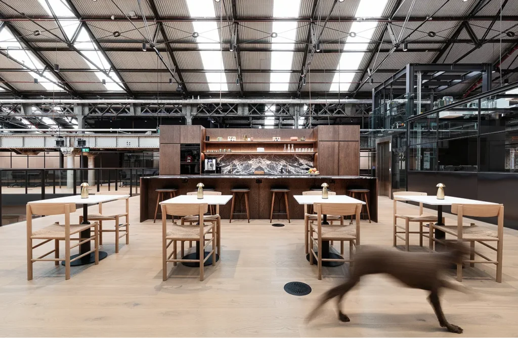 A motion-blurred dog runs across light wood flooring past small dining tables toward a sleek kitchen bar, set within a spacious industrial building featuring high steel trusses and skylights.