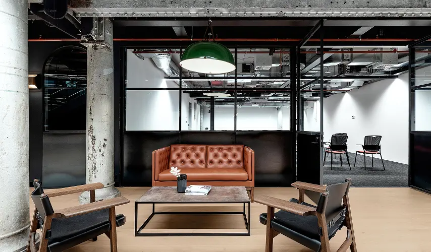 A brown tufted leather sofa and two black armchairs surround a coffee table beneath a green dome pendant lamp inside a modern industrial office with glass partitions and concrete pillars.