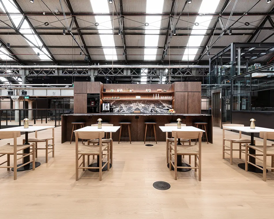 Wooden dining tables with woven chairs sit arranged on light wood flooring before a dark bar counter inside a spacious industrial warehouse with high vaulted skylights.