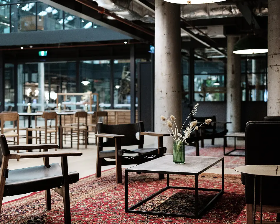Modern black leather armchairs surround a low coffee table holding dried flowers, arranged on a red patterned rug inside a spacious, industrial-style lounge with concrete pillars.