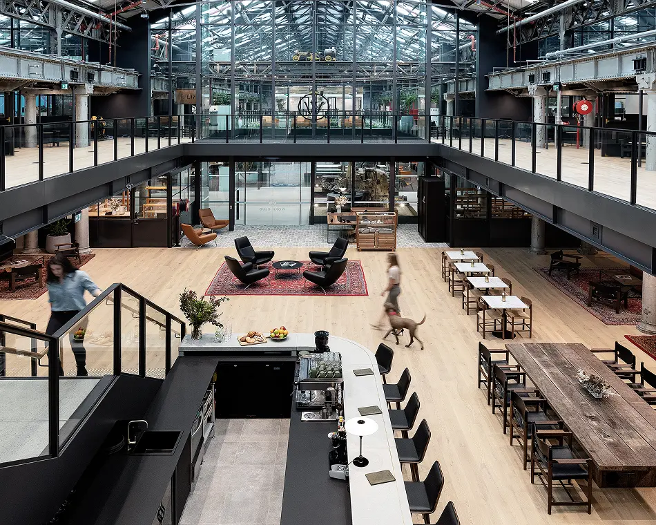 A woman descends stairs near a coffee bar while another walks a dog across a multi-level, light-filled atrium with wooden flooring, seating, and a glass ceiling. Text reads: "WORK CLUB".