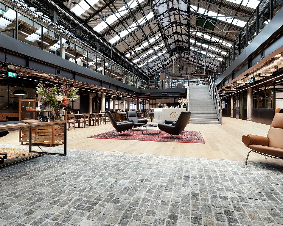 Modern black armchairs surround a coffee table on a red rug, creating a seating area inside a vast, industrial-style atrium featuring light wood floors, a wide staircase, and skylit ceiling.