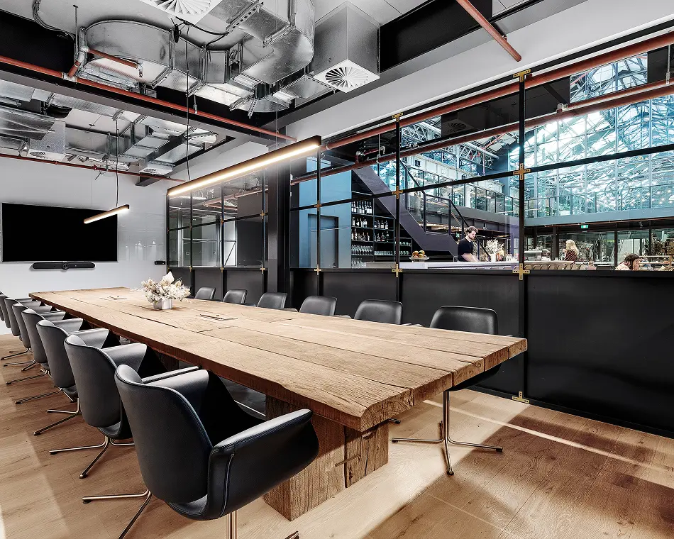 A long, rustic wooden conference table flanked by black leather swivel chairs sits centered in a modern boardroom beneath linear pendant lights, exposed ceiling ducts, and glass-partitioned industrial office space.