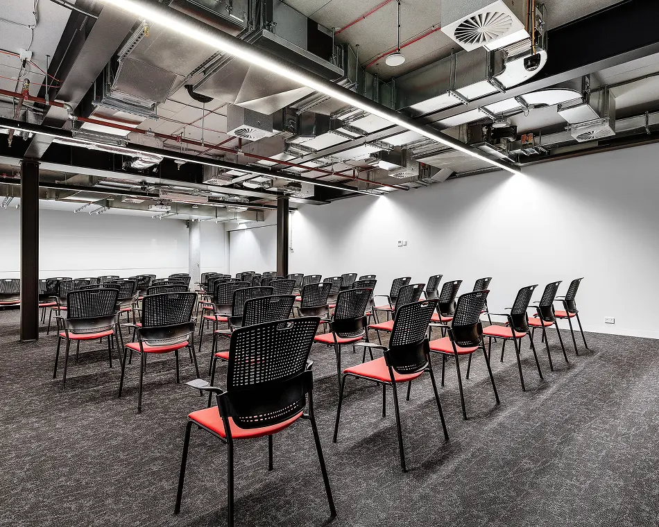 Rows of black mesh-backed chairs with red cushions face forward inside a spacious conference room featuring exposed industrial ceiling ductwork, long linear lights, white walls, and dark carpeting.