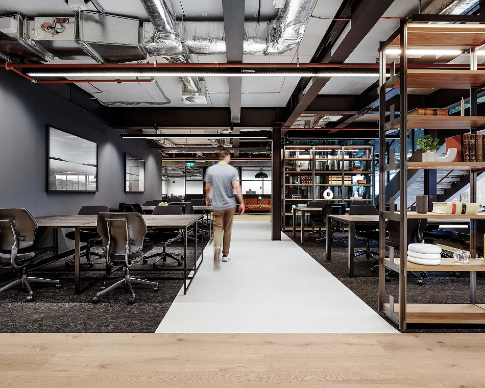 A man walks down a central walkway inside a modern, industrial-style office space, surrounded by dark wooden desks, office chairs, exposed ceiling pipes, and tall open bookshelves.