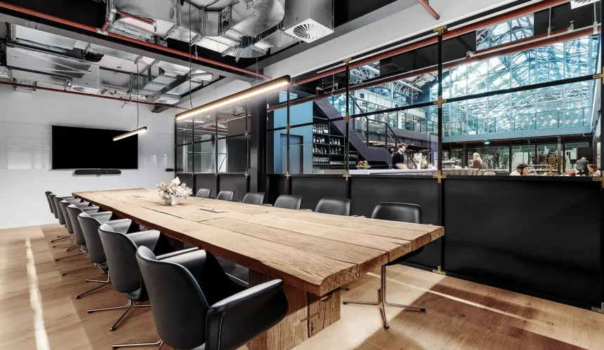 A long, rustic wooden conference table surrounded by black leather chairs sits inside a modern office space featuring exposed ceiling ductwork, a wall-mounted TV, and glass partition walls.
