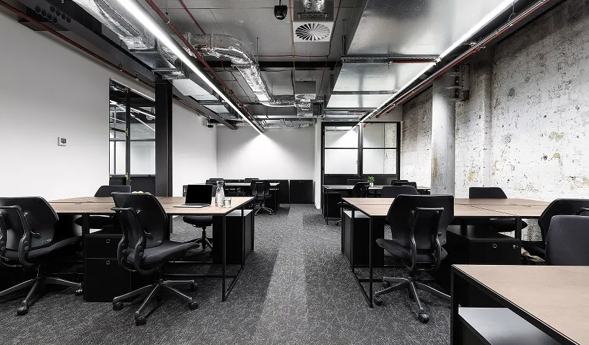 Modern wooden desks paired with black ergonomic chairs sit arranged in an industrial office featuring an exposed ceiling with air ducts, bright linear lighting, and textured concrete walls.