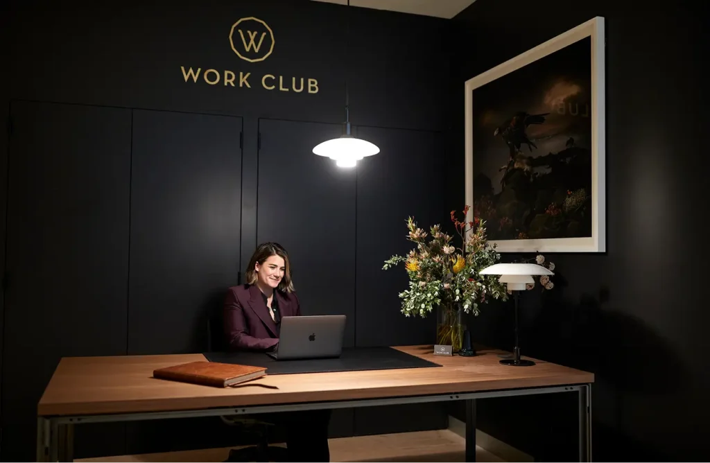 A woman wearing a dark purple blazer sits at a wooden desk working on a laptop inside a dimly lit, black-walled office featuring a floral arrangement and wall logo reading "W WORK CLUB".