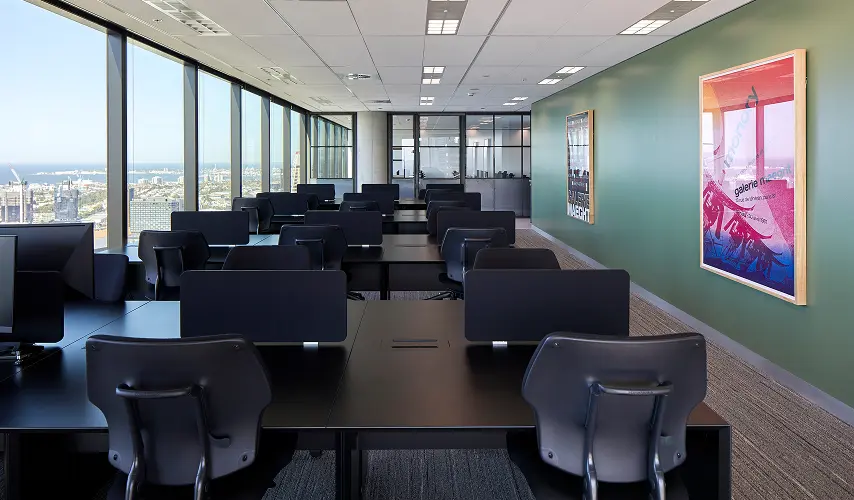 Empty black office desks with computer monitors and ergonomic chairs stand in neat rows within a high-rise office. Floor-to-ceiling windows offer a panoramic coastal view beside a green wall featuring framed posters displaying text: "GALERIE MAEGHT", "monory", "galerie maeght", "13 rue de téhéran, paris 8", and "mai - juin 1971".