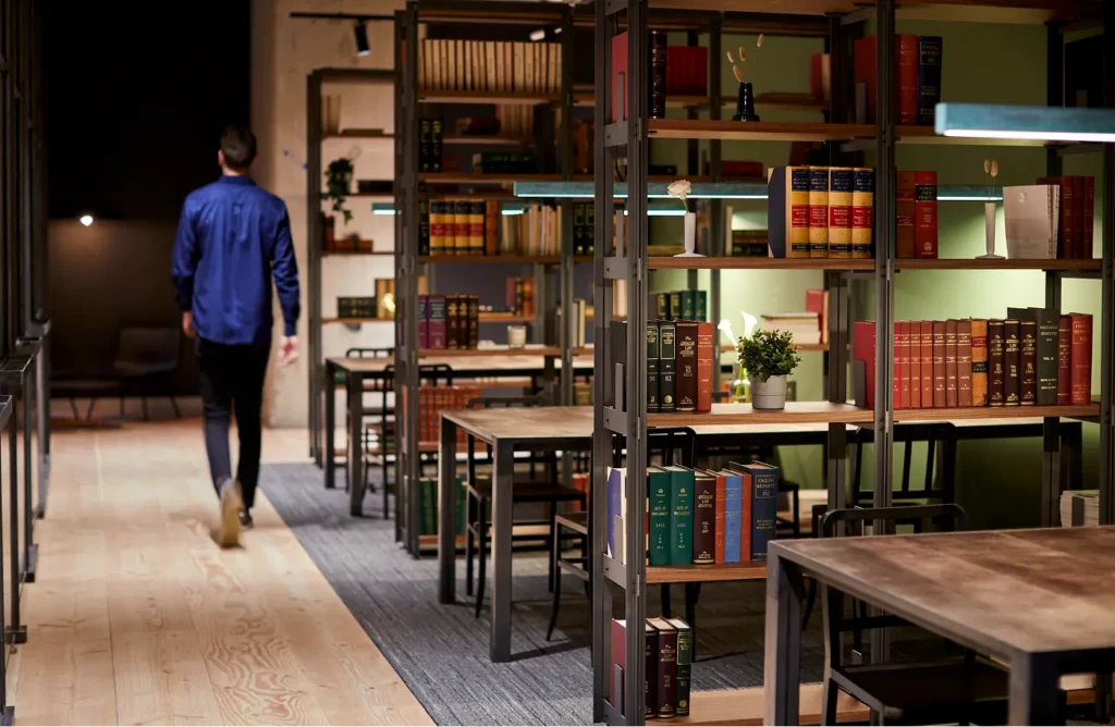 A man wearing a blue shirt walks away down a wooden path inside a warm library lined with tall book-filled shelves, study tables, and chairs. Text on book spines includes "AUSTRALIAN LAW JOURNAL", "ACTS OF PARLIAMENT", "SUPREME COURT PRACTICE", and "ENGLISH REPORTS".