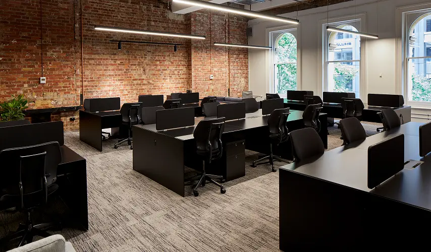 Sleek black office desks paired with ergonomic chairs fill an open-plan workspace, set against an exposed red-brick wall, suspended linear lights, patterned carpet, and large arched windows.