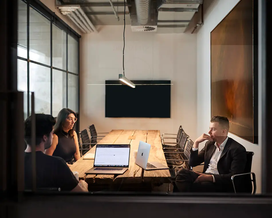 Three colleagues hold a meeting around a long rustic wooden table with open laptops, inside a modern, glass-walled conference room featuring an exposed industrial ceiling.