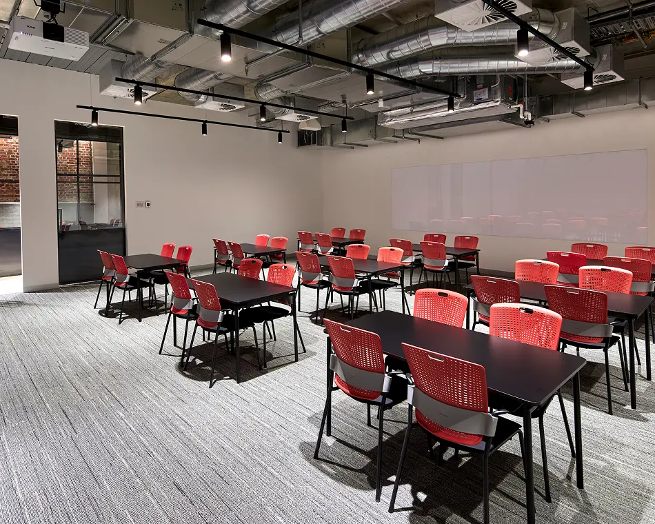 Black rectangular tables paired with bright red mesh chairs sit neatly arranged throughout a modern classroom featuring patterned grey carpet, plain white walls, a whiteboard, and exposed ceiling pipes. The word "EPSON" is printed on the projector.