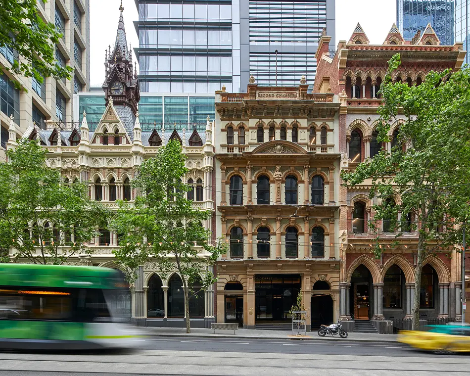A green tram speeds past ornate, Victorian-era historic buildings lined with trees along a city street, framed by modern skyscrapers behind them. Text on the central building reads: “RECORD CHAMBERS” and “OLDERFLEET”.