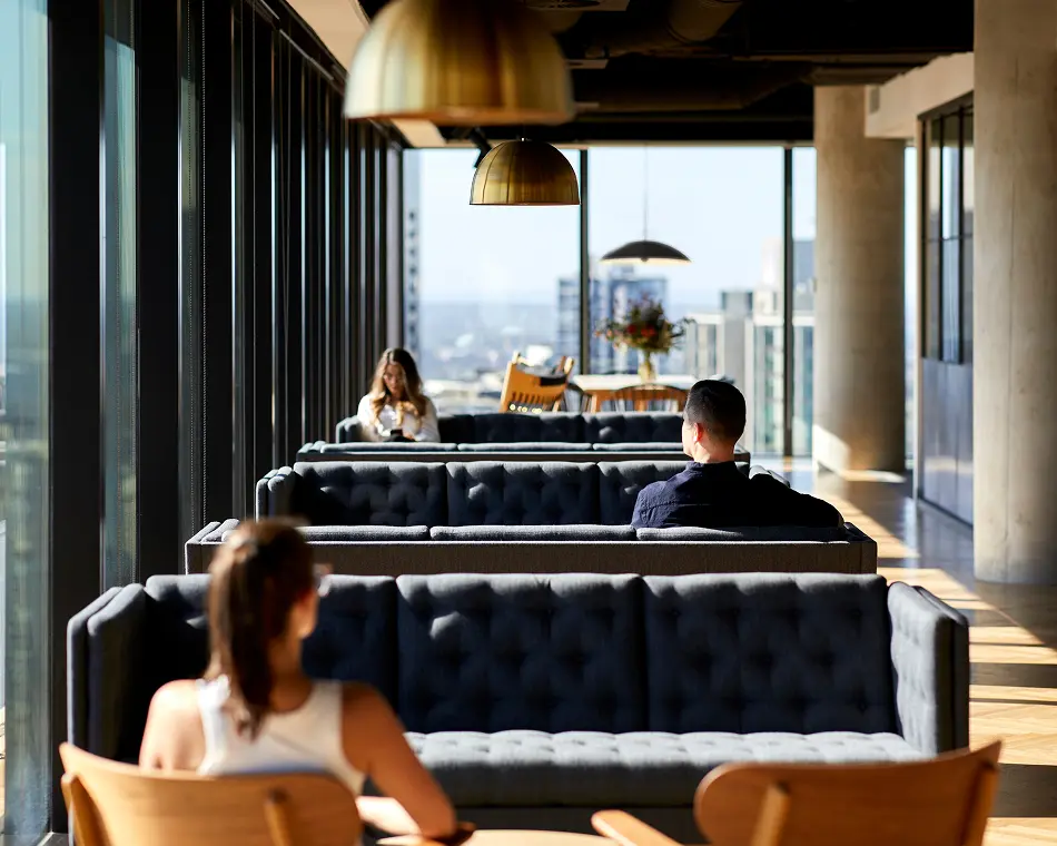 Several people sit and relax on dark tufted couches in a high-rise lounge with floor-to-ceiling windows overlooking a distant city skyline under brass pendant lights.