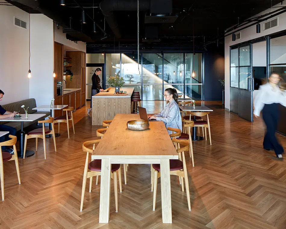 A woman works on a laptop at a long wooden table inside a spacious co-working lounge featuring herringbone timber floors, booth seating, and hanging pendant lights.