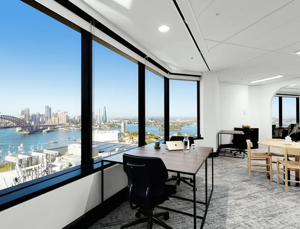 Modern office desks and chairs sit beside large floor-to-ceiling windows, offering panoramic high-rise views of a coastal city skyline, harbor, and iconic bridge under a clear sky.