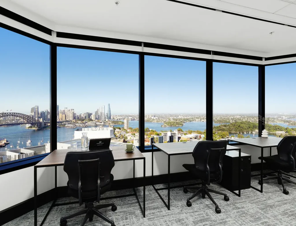 Three black office desks paired with chairs face expansive floor-to-ceiling windows, offering panoramic views of Sydney Harbour and the city skyline. A laptop screen displays text reading "WORK CLUB".