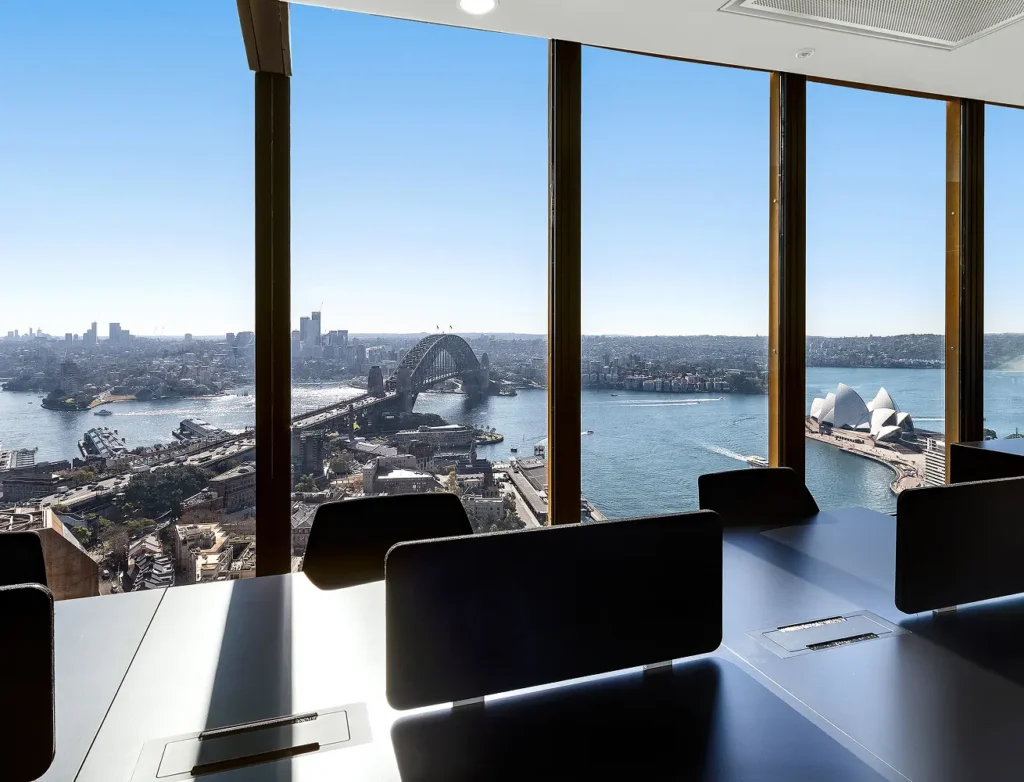 Modern conference desks with black dividers sit inside a high-rise office, offering panoramic views through floor-to-ceiling windows of the Sydney Harbour Bridge, Opera House, and harbour under a bright sky.