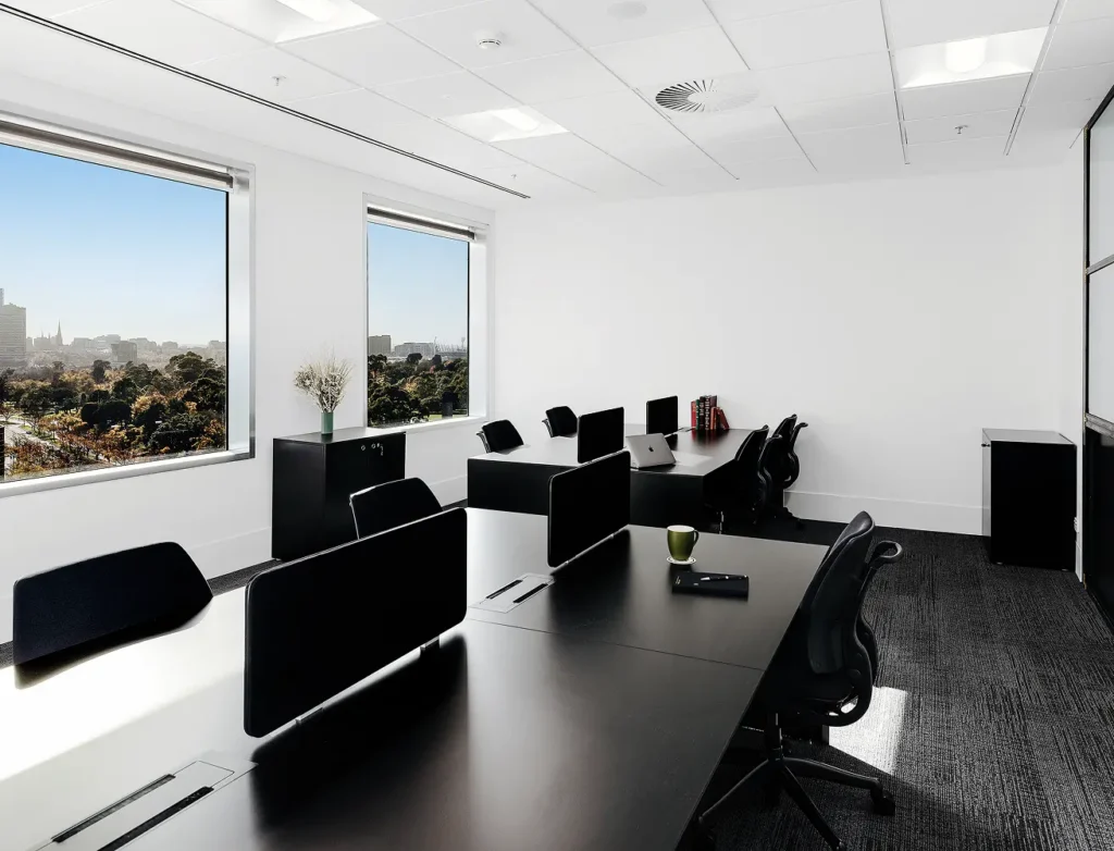 Sleek black office desks equipped with privacy screens and chairs stand organized inside a modern workspace featuring bright white walls, dark carpet, and large windows overlooking city parklands.