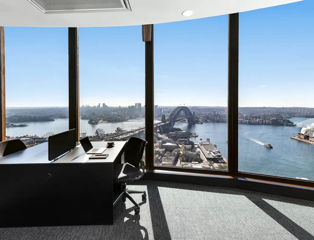 A modern office desk with a laptop sits beside floor-to-ceiling windows, offering a panoramic high-rise view of Sydney Harbour, the Harbour Bridge, and the Opera House on a clear day.