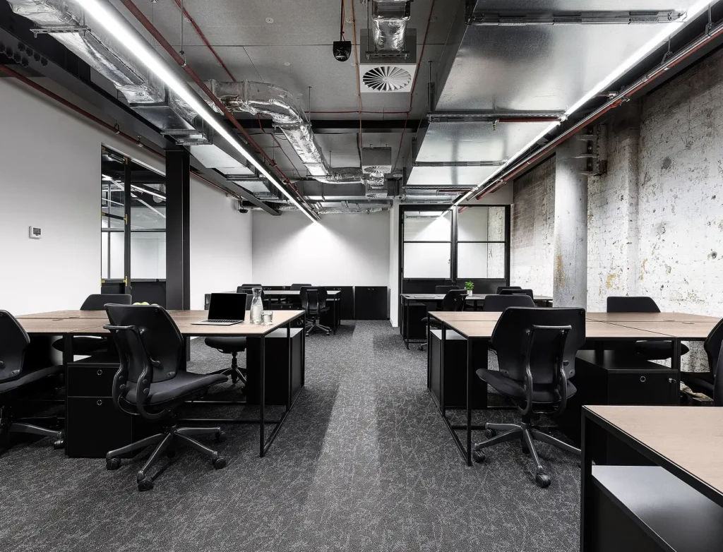 Wooden desks paired with black office chairs sit neatly across a carpeted open-plan room, featuring industrial exposed ceiling ductwork, long strip lighting, and a distressed brick wall.