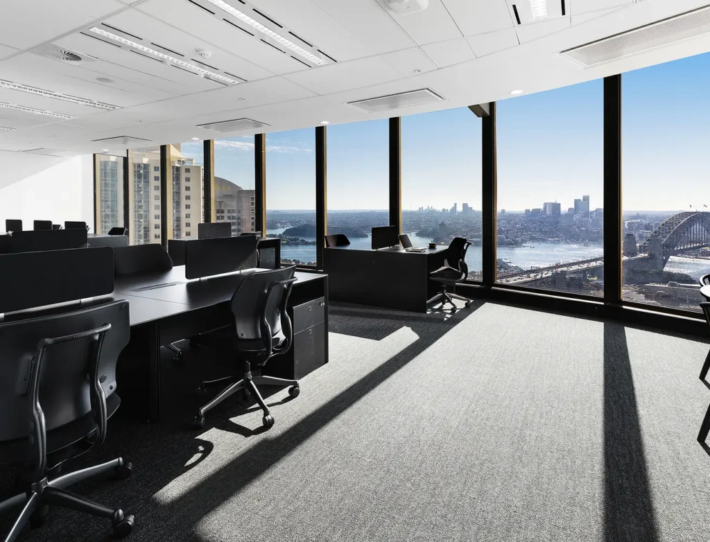Black office desks and ergonomic chairs fill a modern high-rise workspace, surrounded by floor-to-ceiling windows overlooking a panoramic city skyline and harbor under a clear sky.