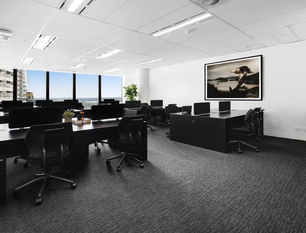 Modern black office desks with monitors and ergonomic chairs sit neatly arranged inside a spacious, carpeted office featuring panoramic city windows and a large framed nature artwork on white walls.