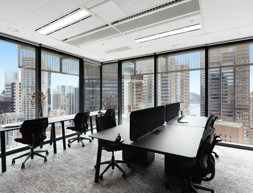 Sleek black office desks and ergonomic chairs sit arranged in a high-rise modern workspace, surrounded by large floor-to-ceiling windows displaying a sprawling cityscape.