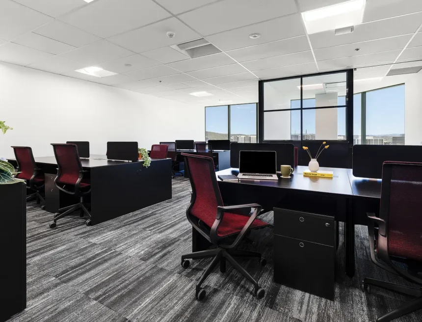 Black workstations paired with burgundy mesh chairs are arranged in rows throughout a modern office featuring gray carpet, drop ceiling lights, and large windows overlooking a distant skyline. Text on the yellow book reads: "Modernist Escapes"