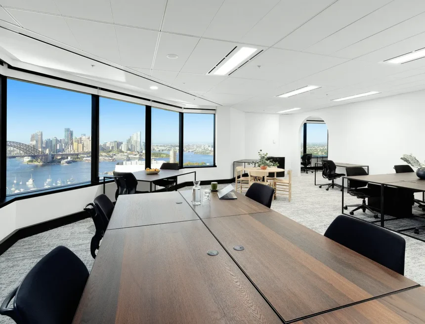 A large wooden conference table surrounded by black chairs fills a modern office space with large panoramic windows overlooking a sunny city harbor skyline.