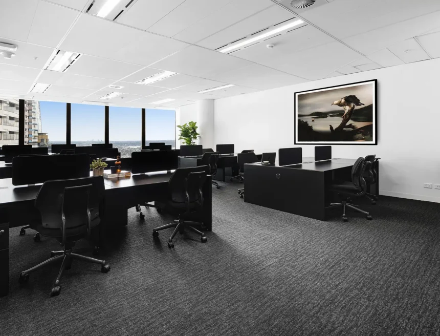 Black modern office desks with chairs and monitors stand arranged across dark carpet inside a high-rise room with large city-view windows and framed eagle artwork.