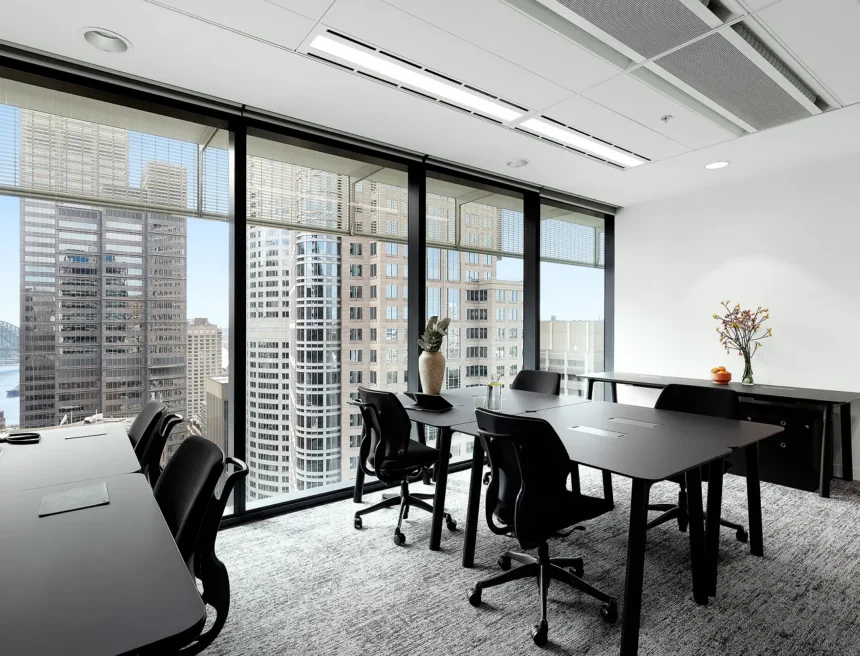 Black office desks and ergonomic rolling chairs furnish a bright, carpeted workspace set against expansive floor-to-ceiling windows that overlook a dense cityscape of skyscrapers.