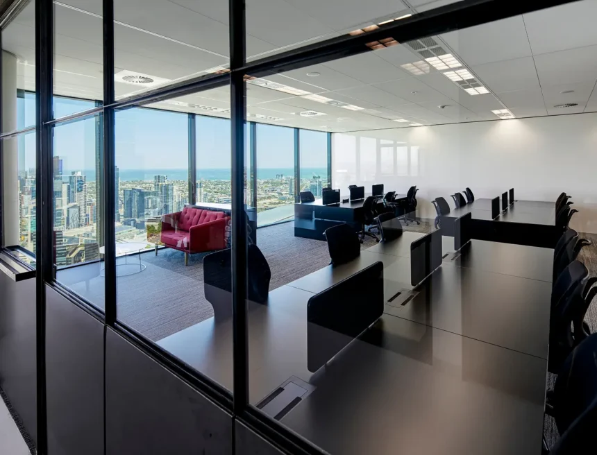 Rows of sleek black office desks sit inside a modern high-rise workspace, viewed through glass partition panels, set against floor-to-ceiling windows offering panoramic city and ocean views.