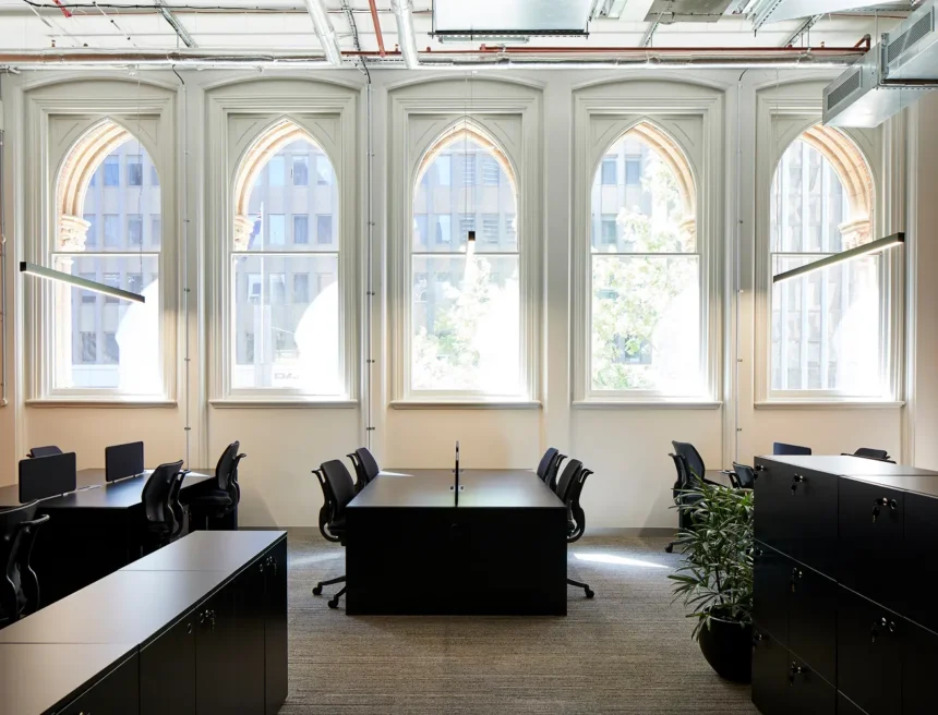 Sleek black office desks and ergonomic chairs stand empty inside a bright workspace featuring five large Gothic-arched windows, exposed ceiling pipes, black storage cabinets, and carpeted flooring.