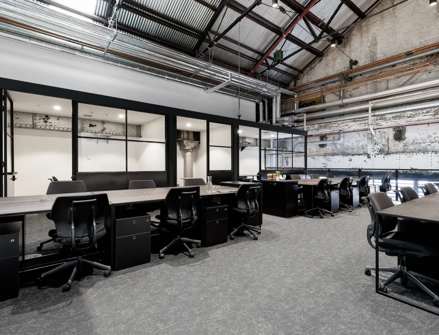 Rows of black office desks and ergonomic chairs stand arranged inside a spacious industrial-style workspace featuring glass-partitioned offices, exposed overhead pipes, high ceilings, and grey carpeted floors.