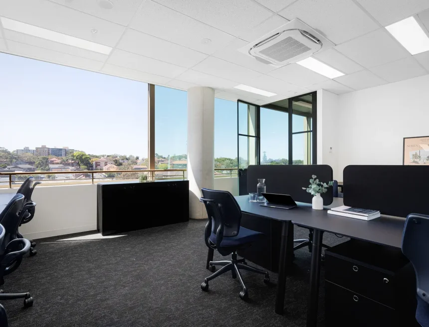 Modern black office desks and navy swivel chairs sit in a bright, carpeted office room featuring large floor-to-ceiling windows overlooking a distant residential landscape under a clear sky.