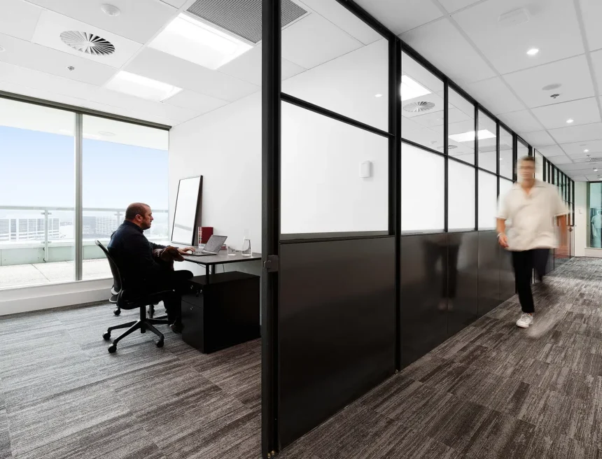 A man works on a laptop at a desk inside a modern office room, while a blurred figure walks along an adjacent carpeted hallway separated by black-framed glass partitions.