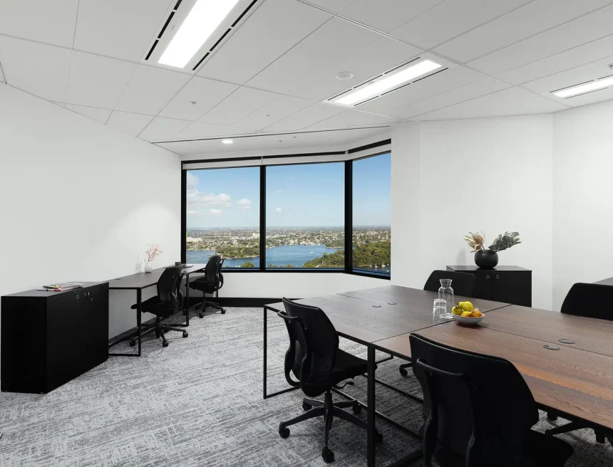 Dark wooden desks paired with black office chairs furnish a bright modern office featuring carpeted floors, overhead lighting, and large bay windows showcasing a panoramic city and harbor view.