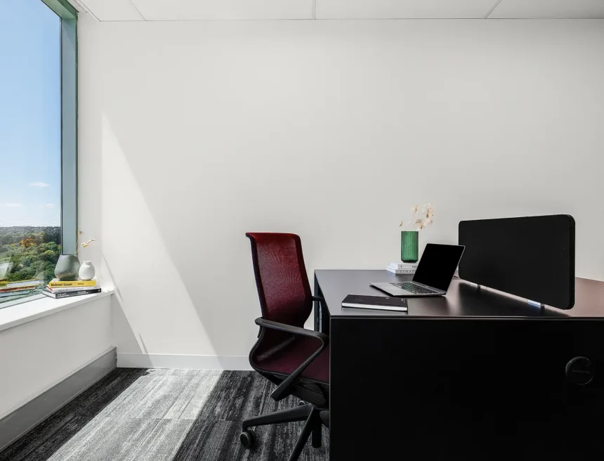 A burgundy mesh chair sits at a dark desk equipped with an open laptop and notebook inside a bright office featuring white walls, striped carpet, and a large window. Text visible on book spines: "Modernist Escapes", "The Icon", "PRINCIPLES OF STYLE", "THE CITY GARDENER", "RICHARD LONGWORTH", "Mein...", "CURATI...".