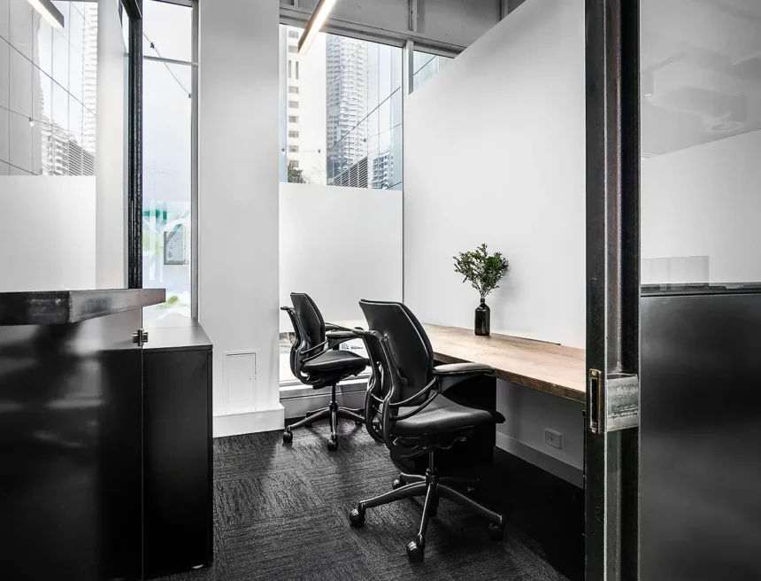 Two black ergonomic office chairs sit at a long wooden desk featuring a small vase, set within a modern office space with large windows overlooking city skyscrapers.
